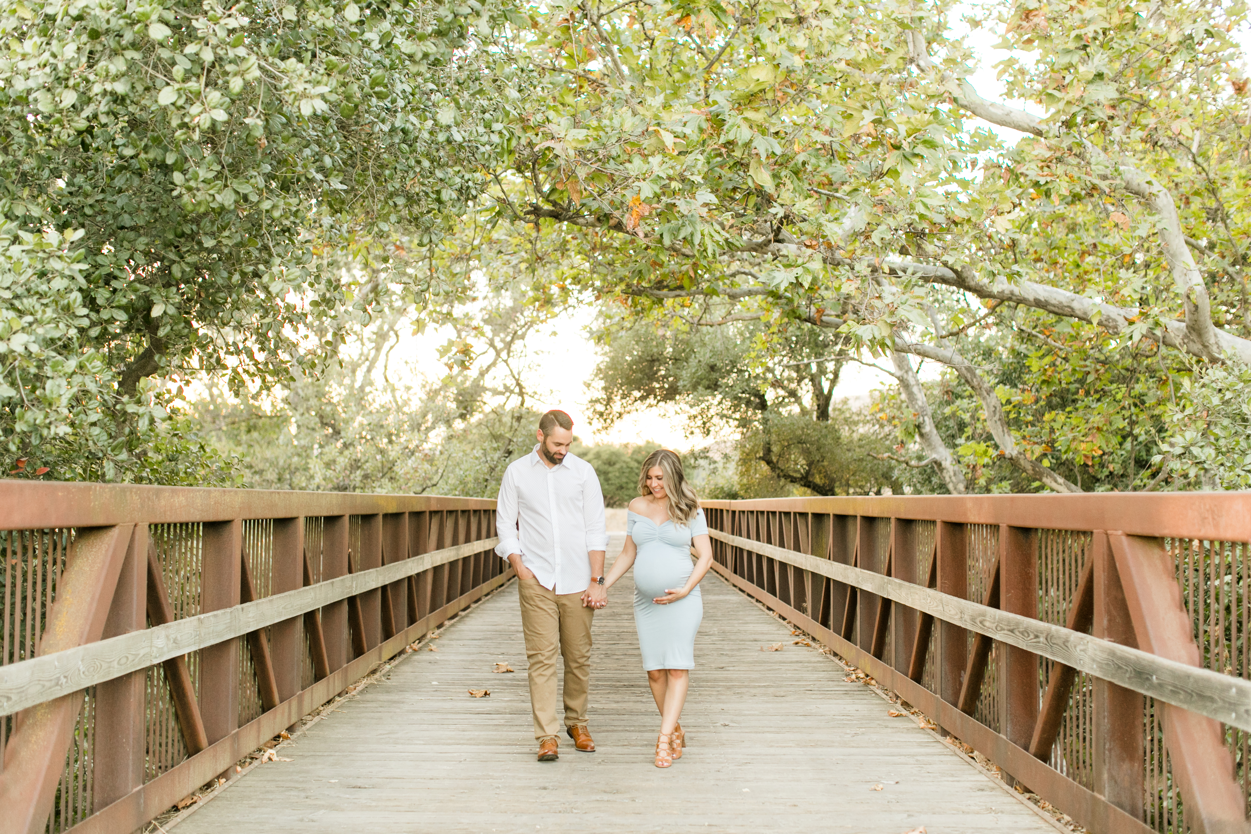 husband and wife walking holding hands across bridge in morgan hill california while mom to be holds belly for maternity photos