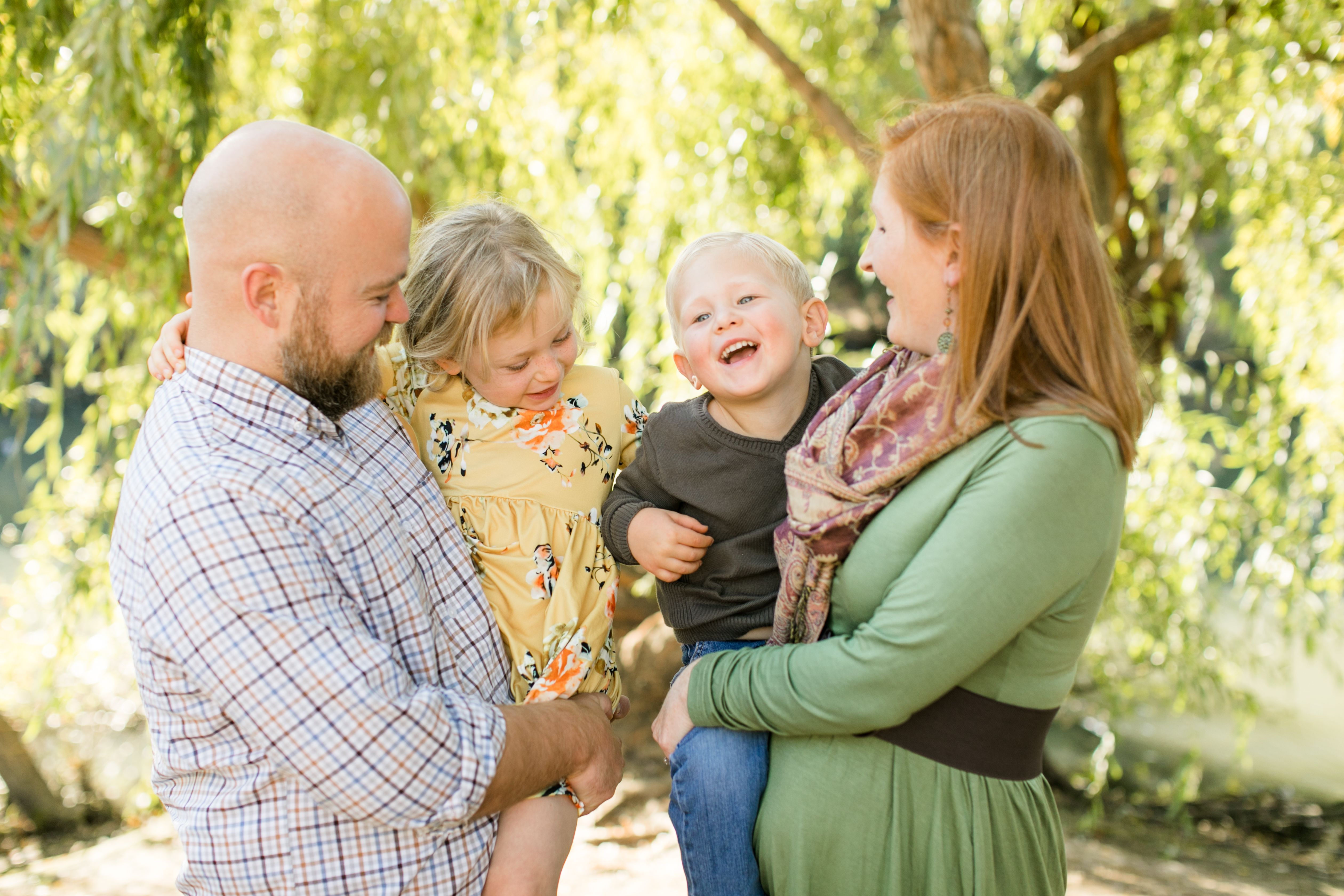 family of four standing in front of tree mom holding young son and dad holding young daughter all laughing at each other