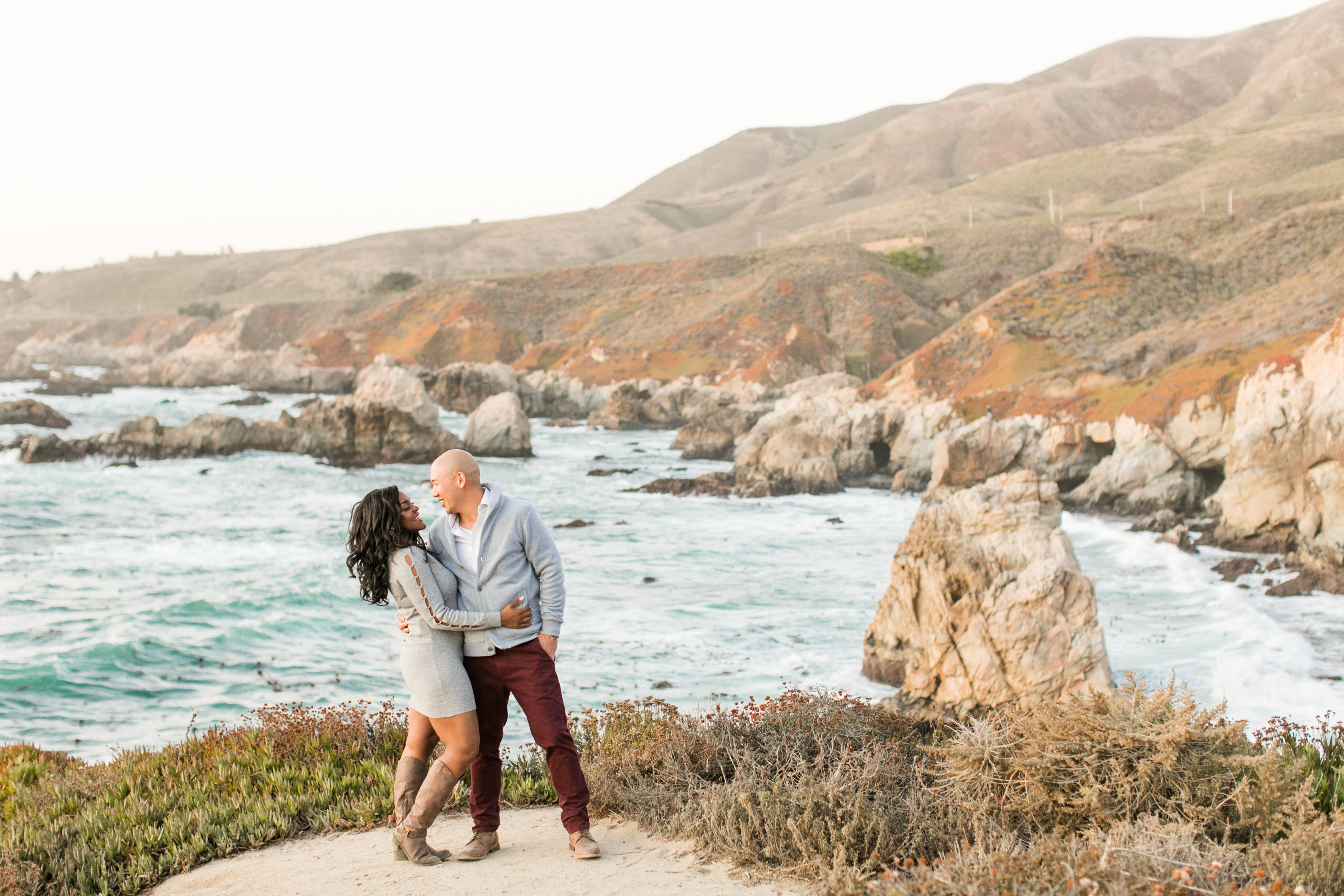 man and woman stand on the cliffs in Carmel overlooking the ocean holding each other in loving embrace and laughing at each other