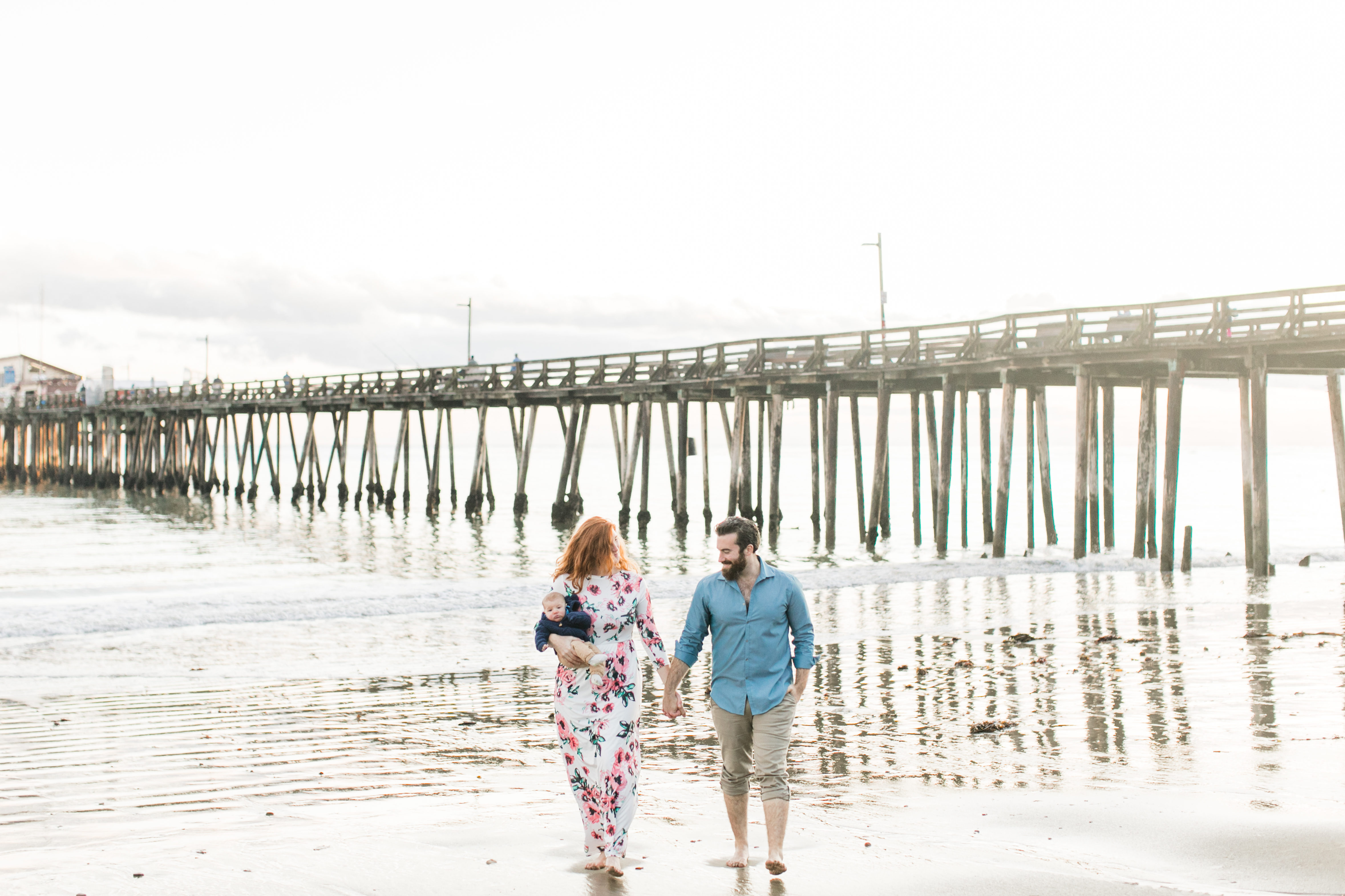 wife and husband walk hand in hand with 3 month old baby in wife's arms on the beach with a the pier behind them