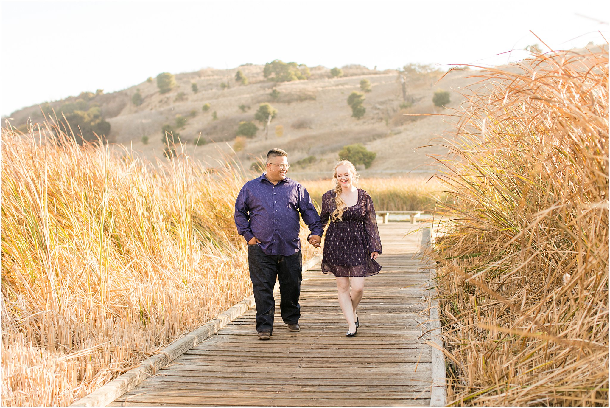 man engaged to woman holding hands walking down dock at golden hour with tall wheat grass around them and hills behind them