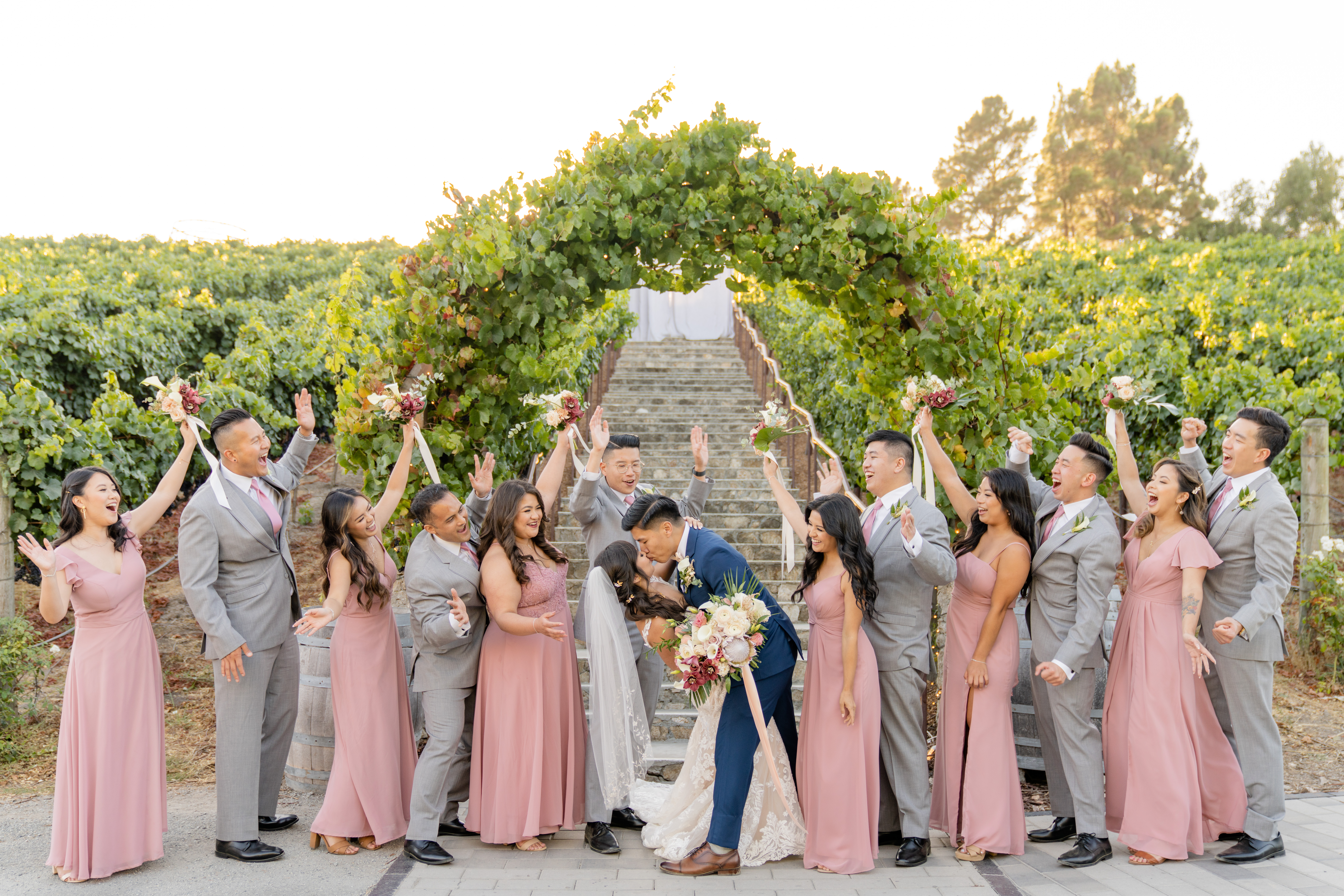 newlyweds share an epic kiss under vineyard arch surrounded by their wedding party cheering them on