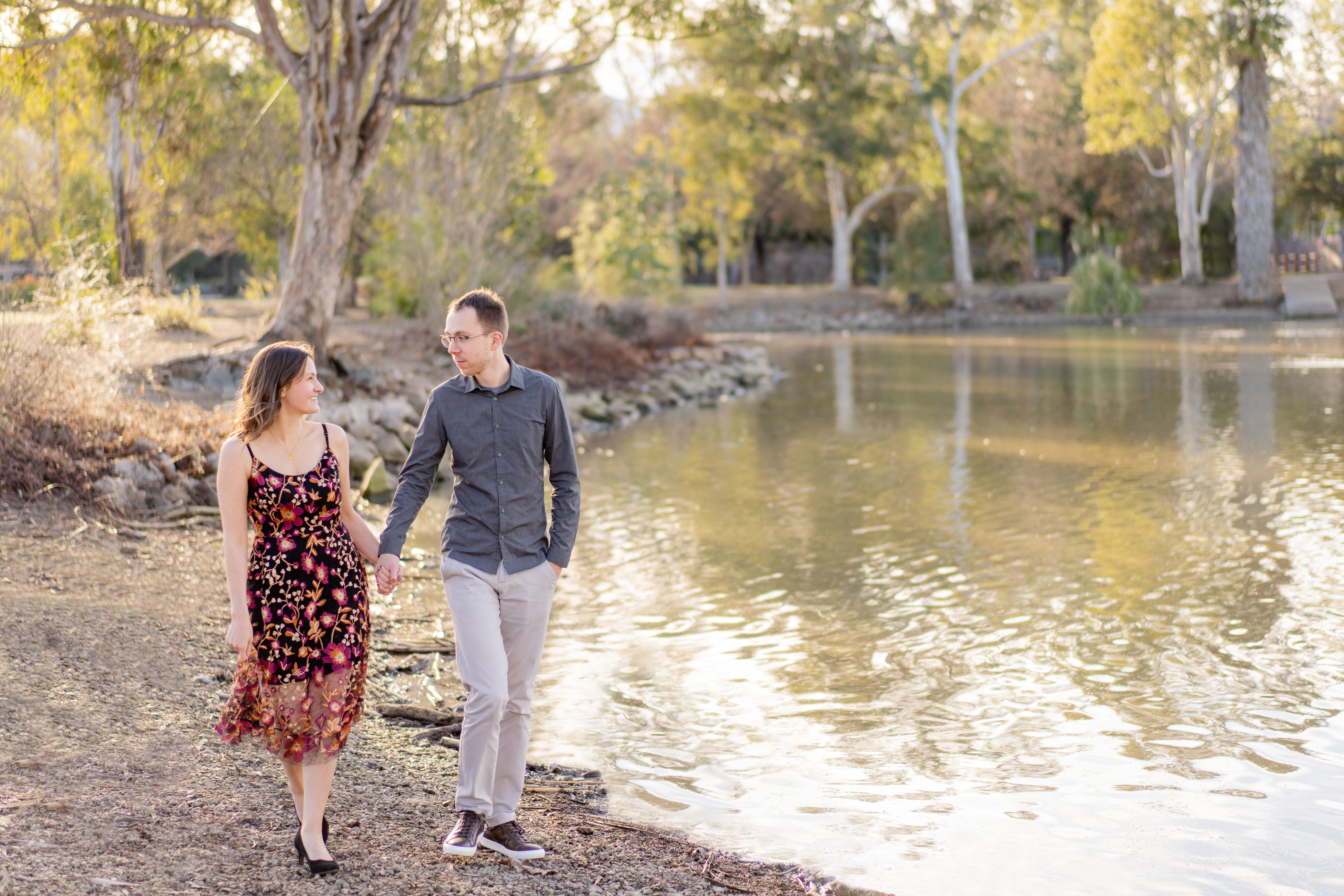 engaged couple holding hands while walking alongside a lake