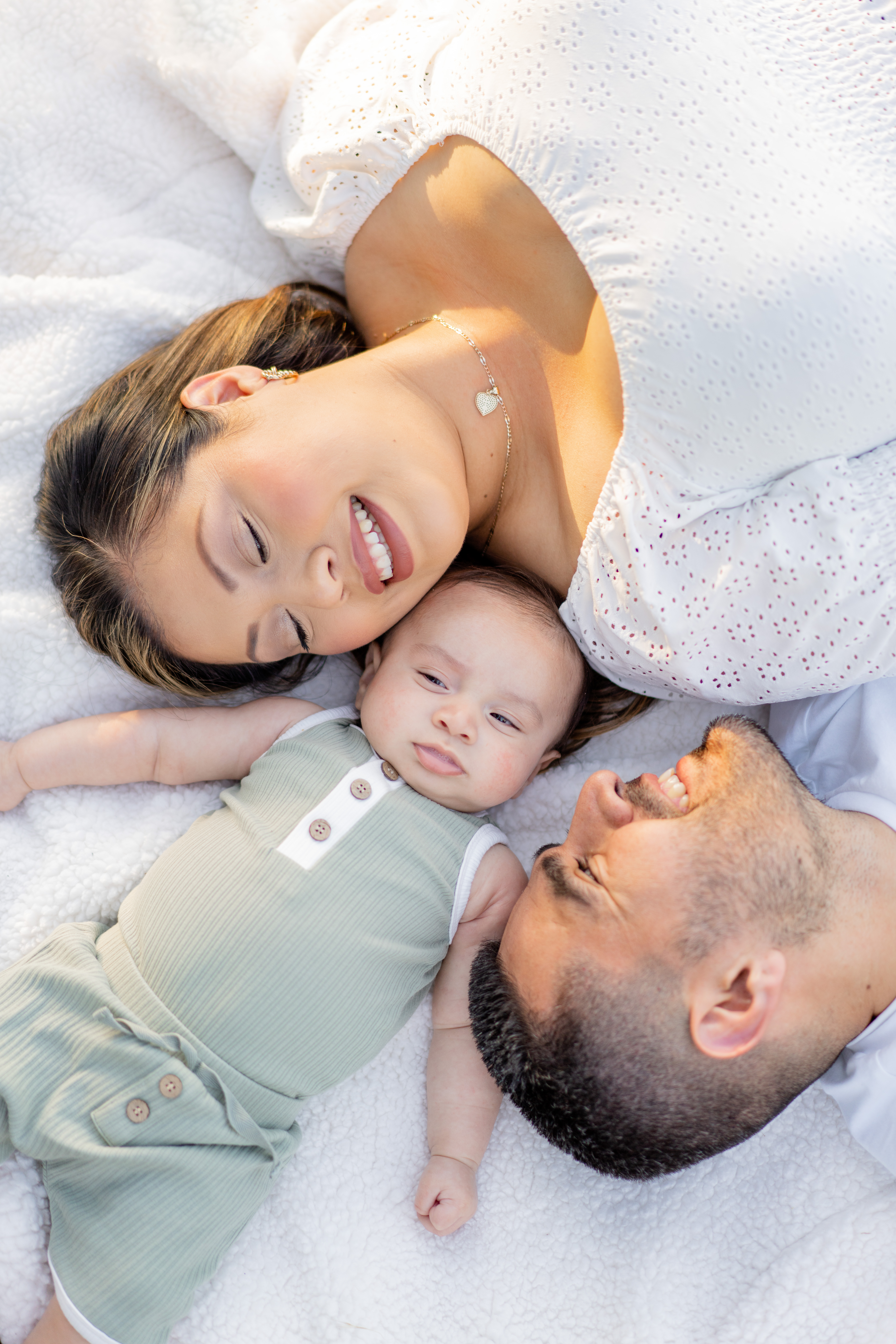young family of three lay down on a blanket together. mom and dad are on either side of baby boy laughing and smiling