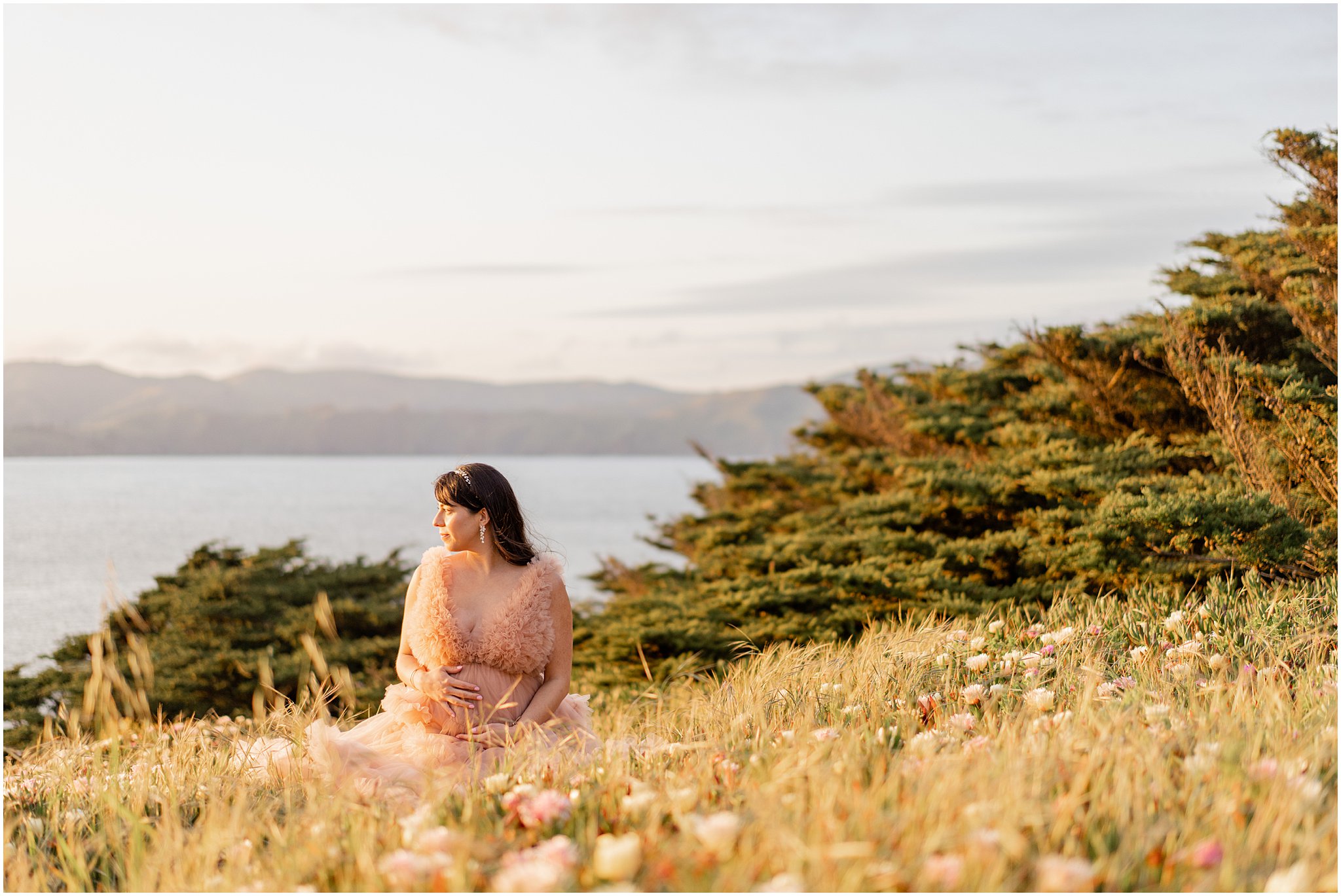 pregnant mom sits wistfully in a field of flowers overlooking the pacific ocean