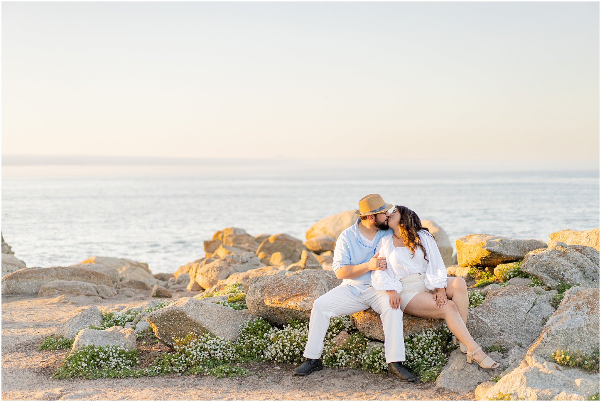 engaged couple sit cuddled on a rock formation by the Pacific Ocean. They share a kiss while the sun sets behind them.