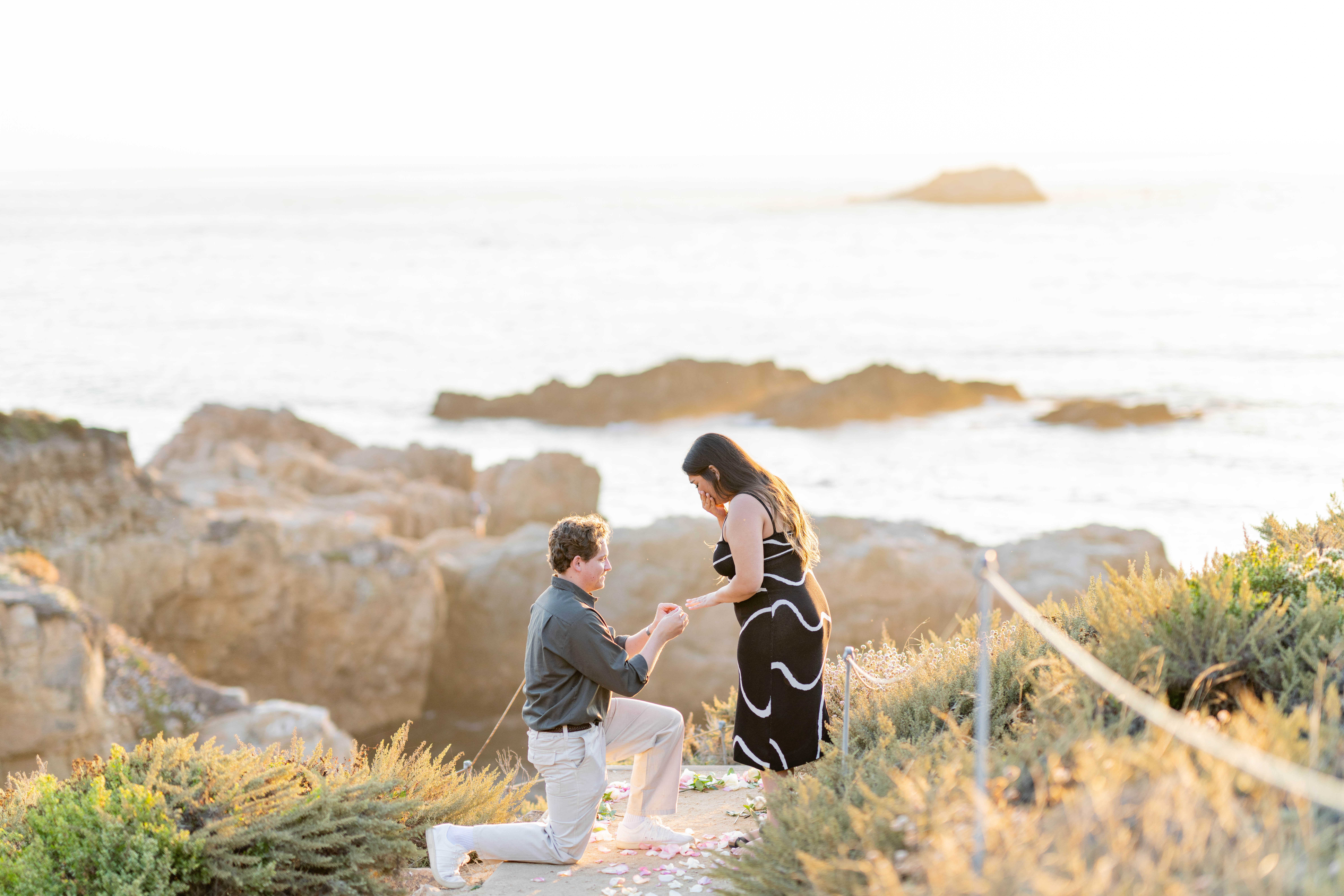 landscape image of young man down on one knee proposing to his girlfriend who is standing - he is sliding the ring on her finger right after she said yes