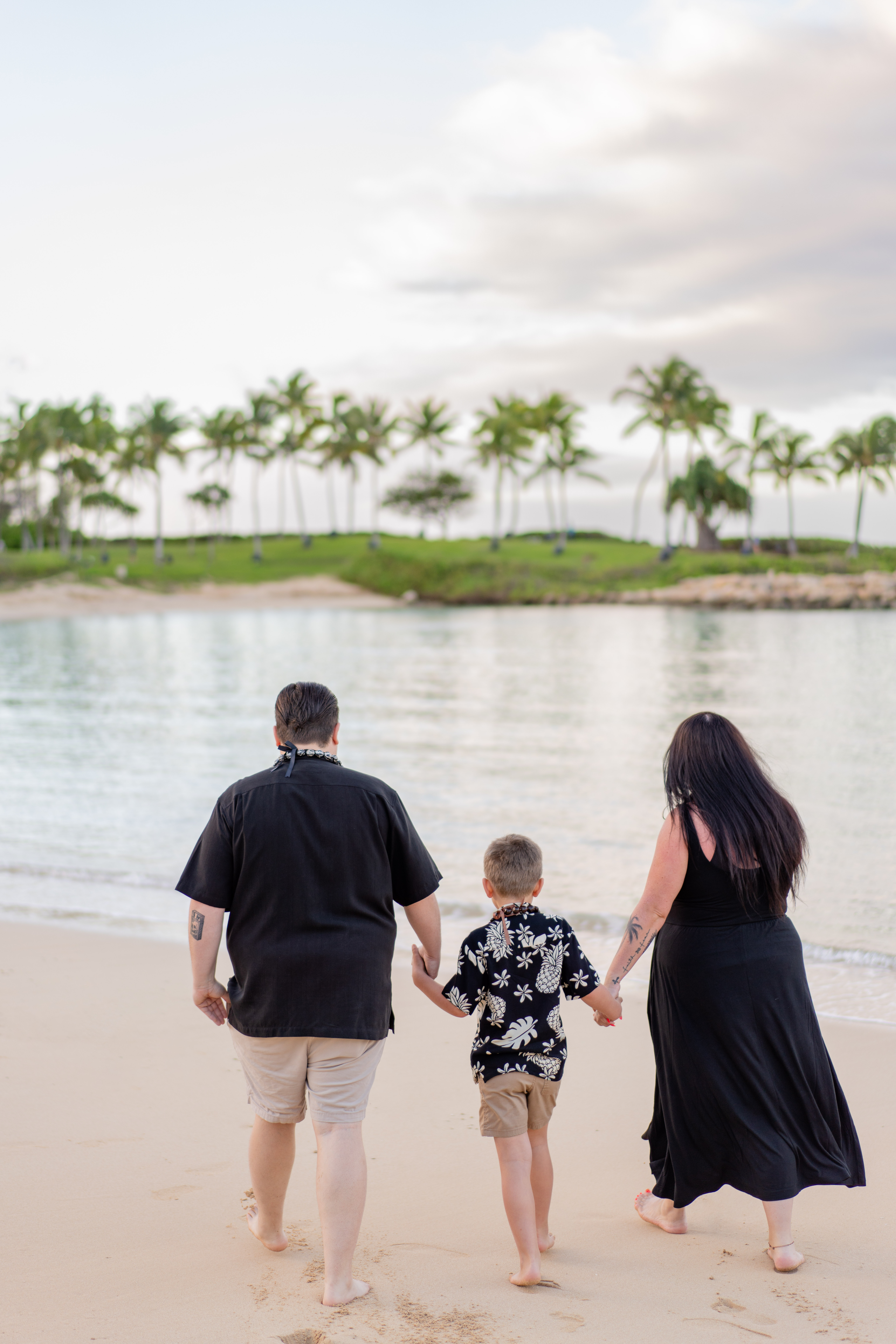 a family of three - dad, mom and young son - walk away from the camera hand in hand on the beach with palm trees and cloudy skies ahead of them