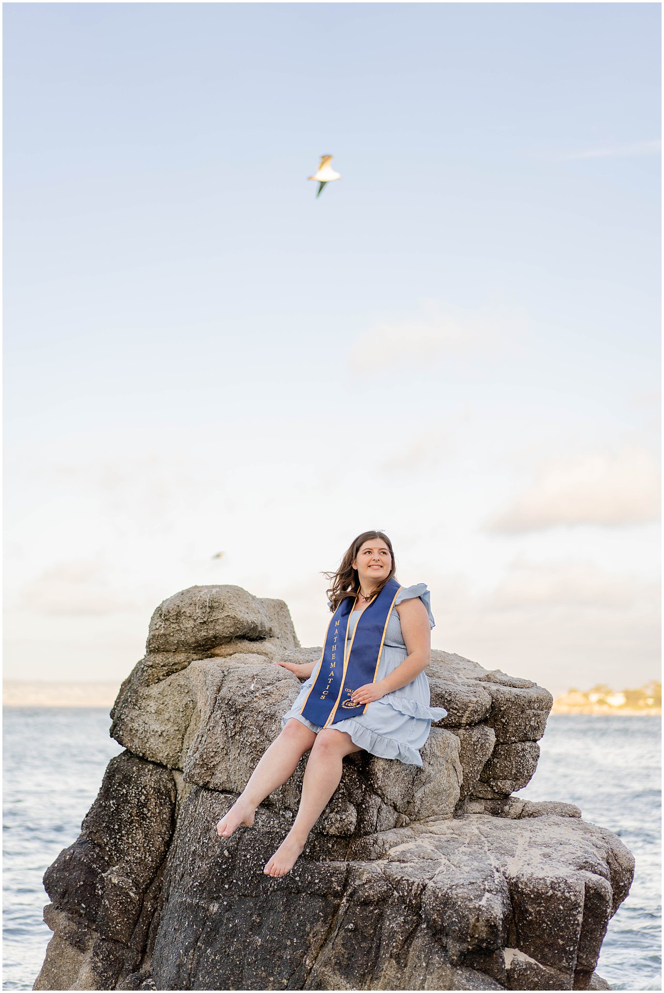 college graduate woman sits on a tall rock overlooking the pacific ocean with seagulls flying above her