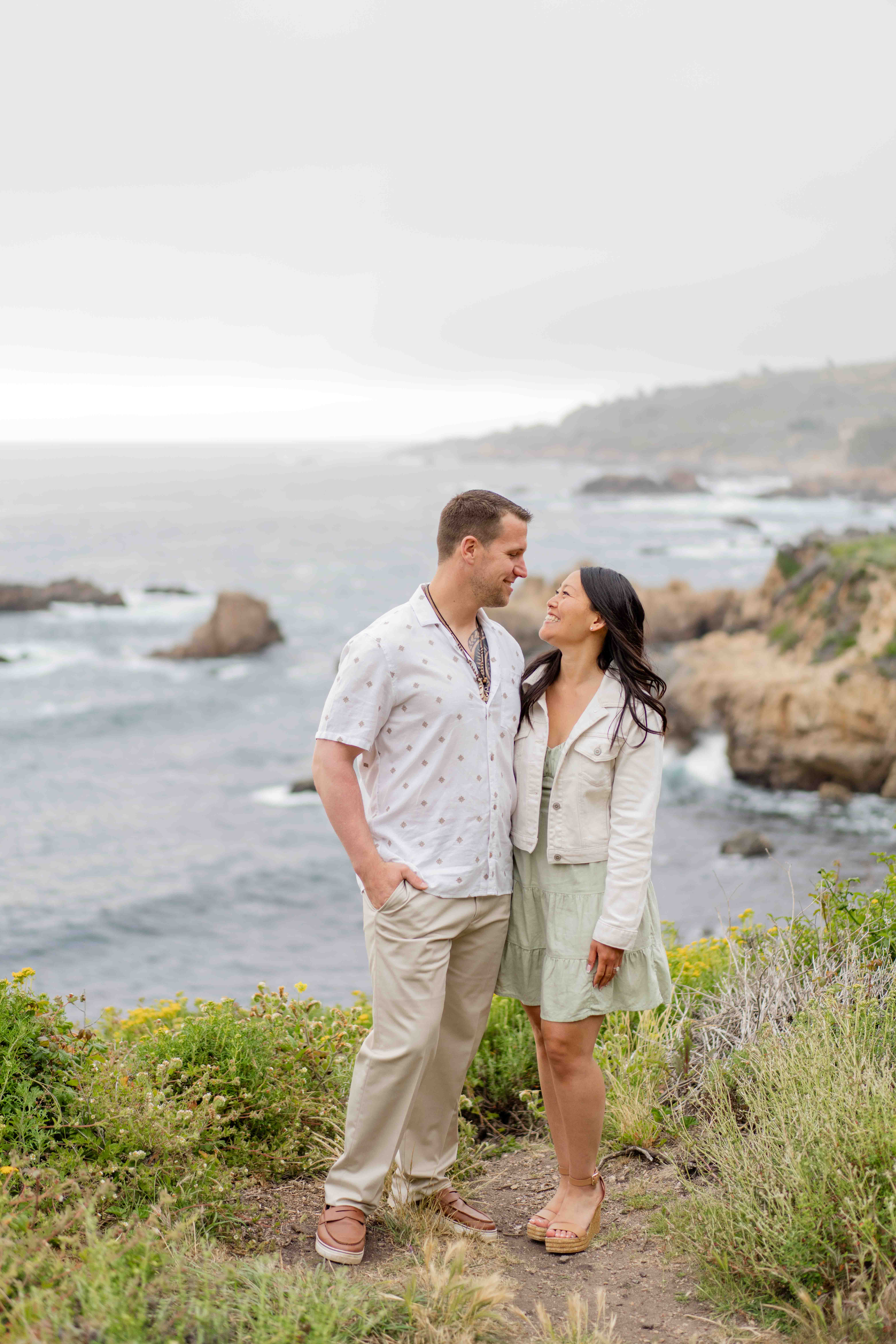 engaged couple standing side by side looking lovingly at each other with the pacific ocean and california cliffs behind them